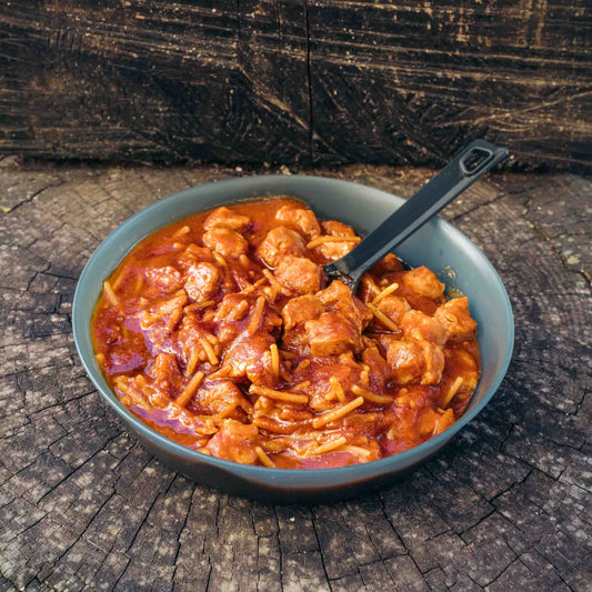 Gulasch Outfood plant based con bocconcini di soia e pasta, servito in una ciotola grigio-blu con cucchiaio nero, su tronco di legno.