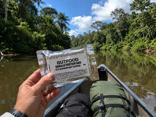 Mano che tiene una barretta proteica Outfood Vanilla da 45g in confezione argentata, su una canoa in un fiume tropicale.