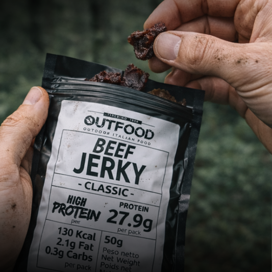 Mani sporche tengono una bustina nera Outfood Beef Jerky Classic da 50g e un pezzo di carne essiccata.