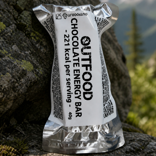 Barretta energetica Outfood Chocolate Energy Bar da 40g, 221 kcal, in confezione argentata e bianca su roccia.