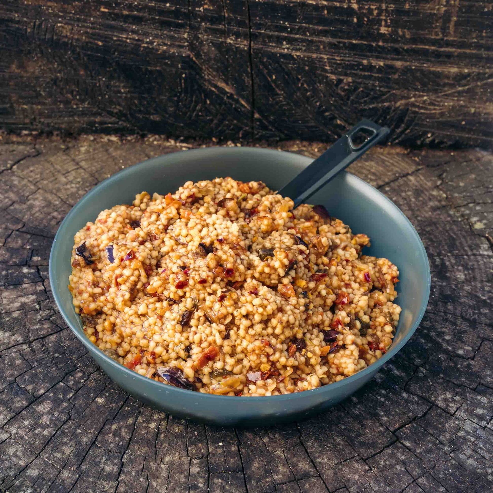 Couscous con caponata di verdure Outfood in ciotola grigia con cucchiaio nero, su superficie di legno scuro.