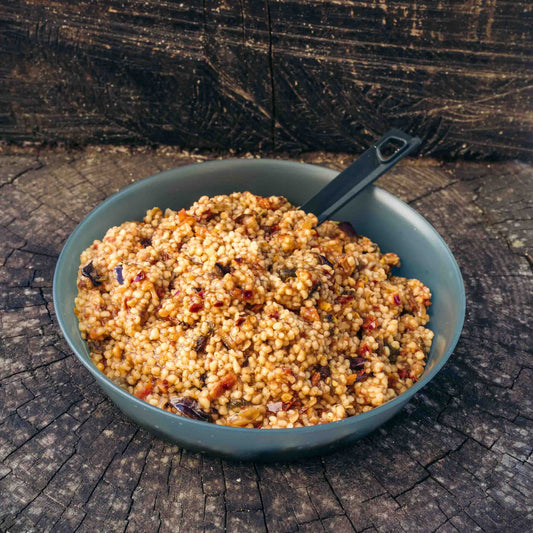 Couscous con caponata di verdure Outfood in ciotola grigia con cucchiaio nero, su superficie di legno scuro.