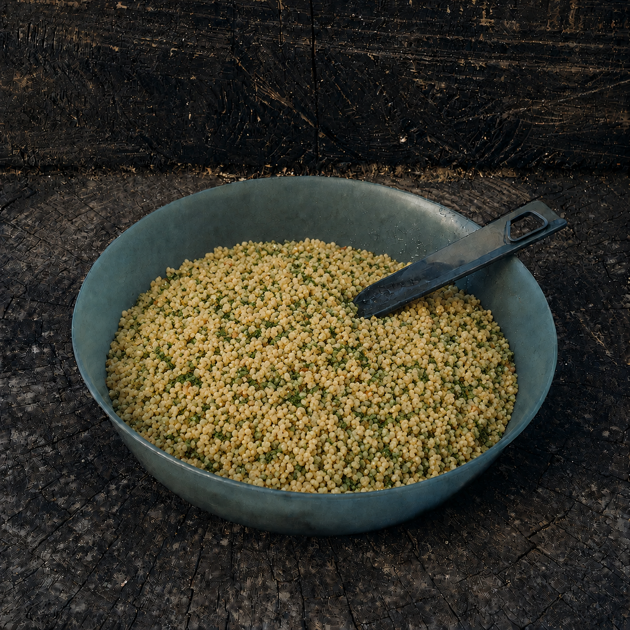 Couscous con erbe fini e spezie Outfood in una ciotola verde scuro con cucchiaio nero, su legno scuro.