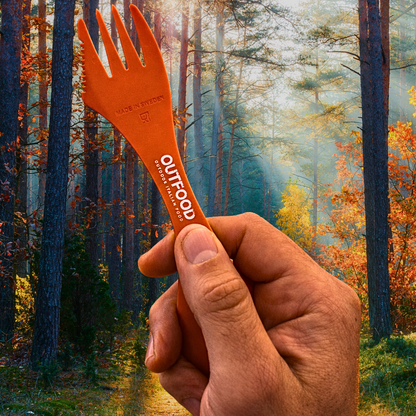 Mano che tiene una spork Outfood arancione con scritta "MADE IN SWEDEN" e "OUTFOOD OUTDOOR ITALIAN FOOD" su sfondo boschivo autunnale.