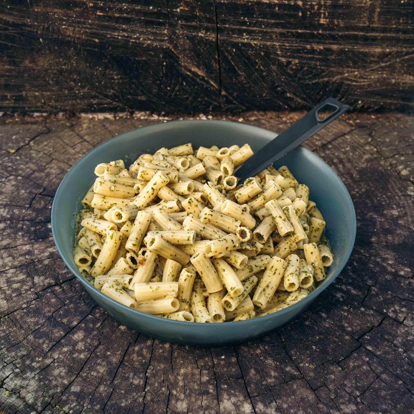 Pasta al pesto di basilico alla genovese Outfood in una ciotola grigia con cucchiaio nero su tronco di legno.
