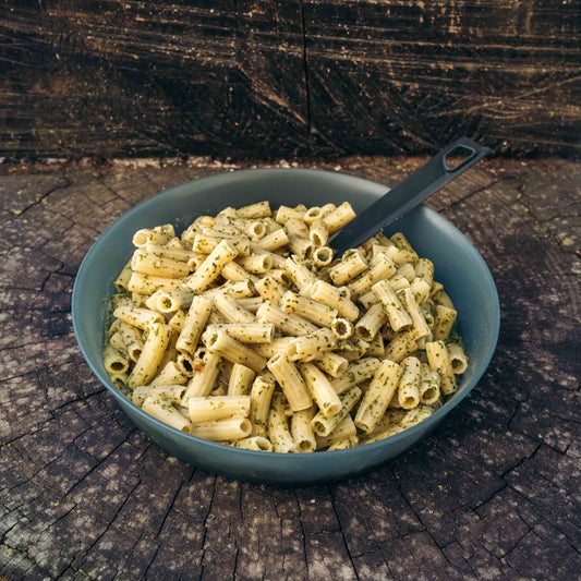 Pasta al pesto di basilico alla genovese Outfood in una ciotola grigia con cucchiaio nero su tronco di legno.