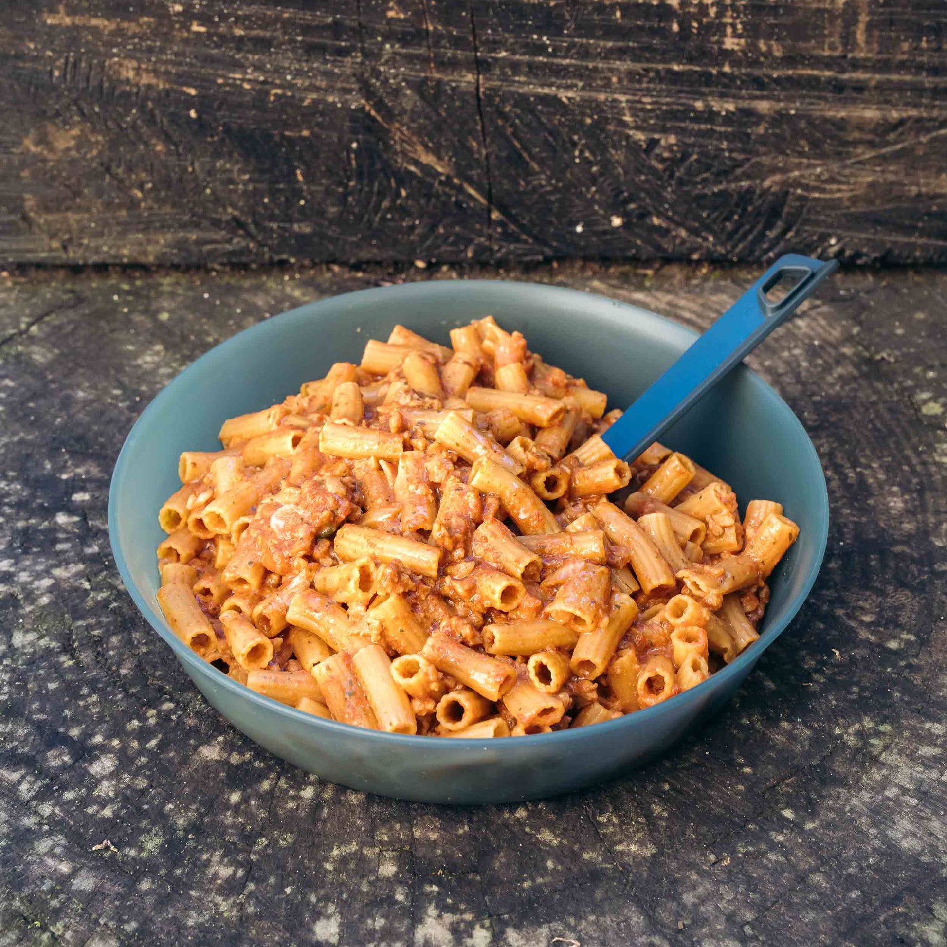 Pasta rigatoni con ragù alla bolognese vegano in ciotola blu scuro con cucchiaio blu, su tronco di legno.
