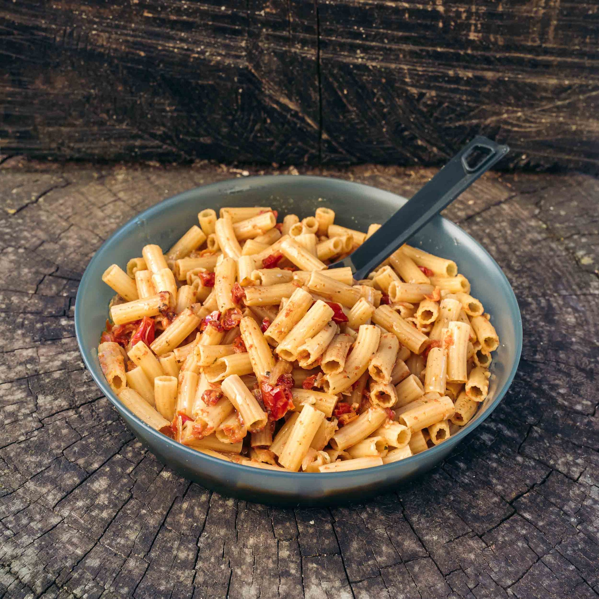 Pasta al sugo di pomodoro e basilico in una ciotola grigia con cucchiaio nero, su un tronco d'albero.