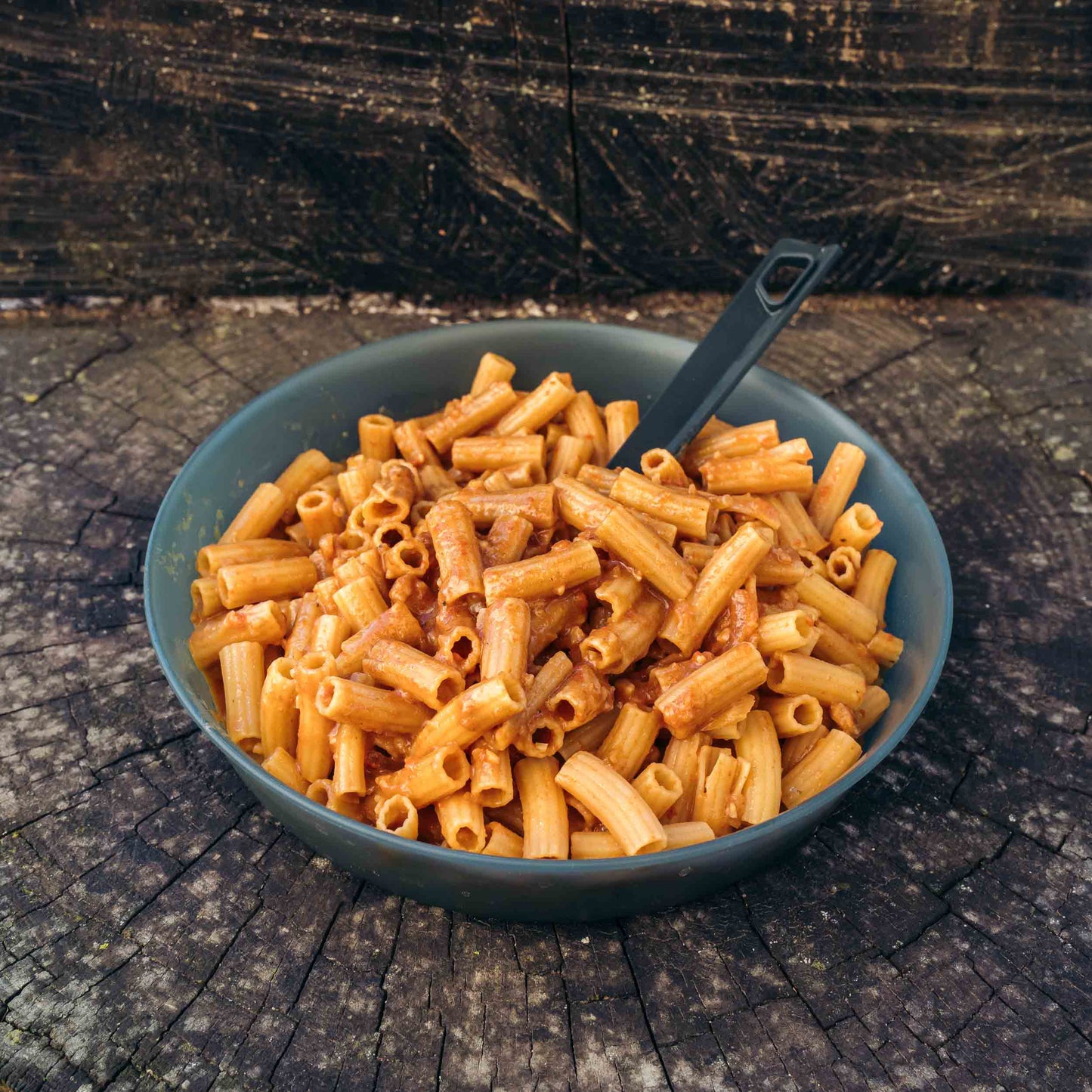 Pasta all'Amatriciana Outfood in ciotola grigia con salsa rossa e cucchiaio nero su tronco di legno scuro.