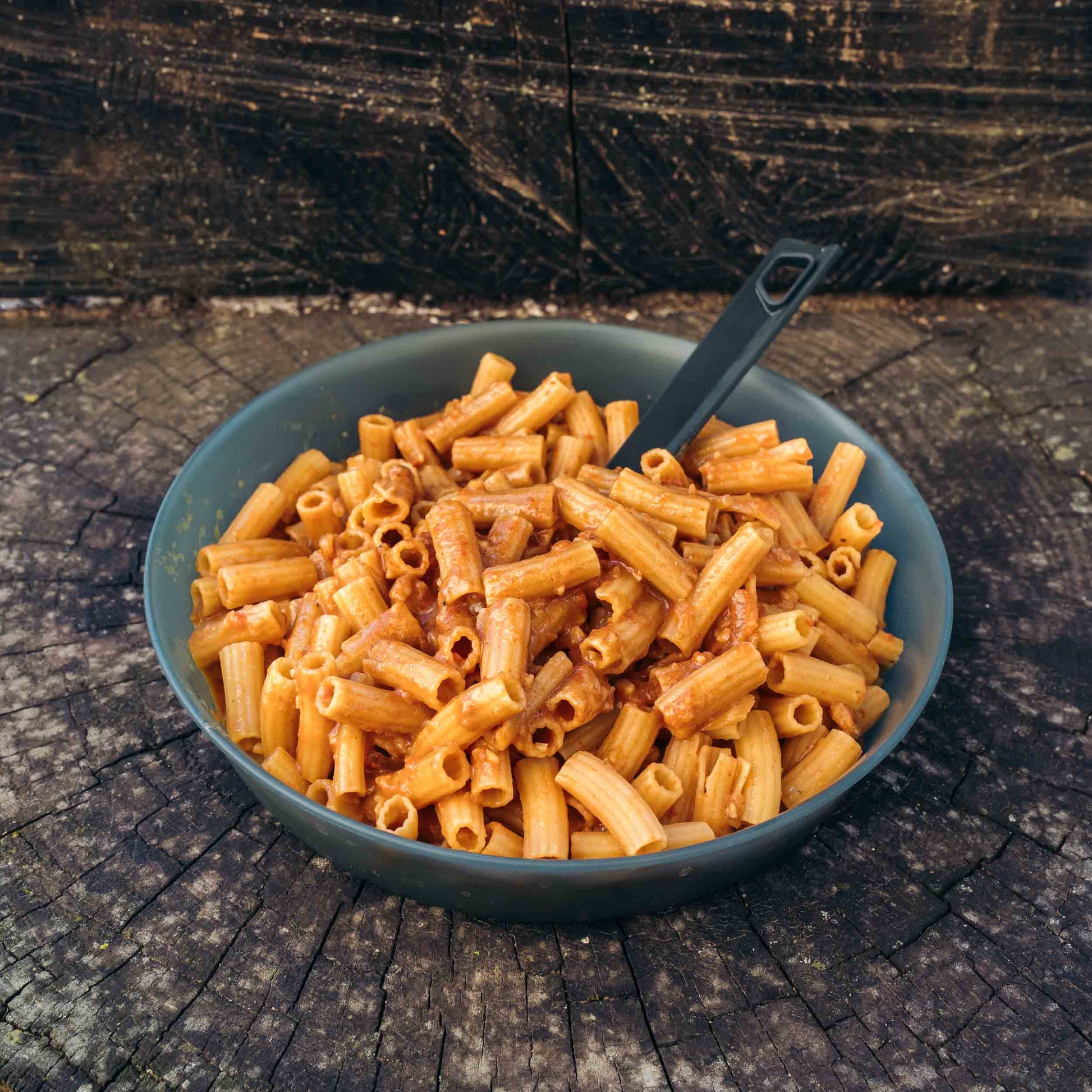 Pasta all'Amatriciana Outfood in ciotola grigia con salsa rossa e cucchiaio nero su tronco di legno scuro.