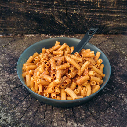 Pasta all'Amatriciana Outfood in ciotola grigia con salsa rossa e cucchiaio nero su tronco di legno scuro.