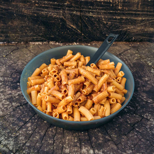 Pasta all'Amatriciana Outfood in ciotola grigia con salsa rossa e cucchiaio nero su tronco di legno scuro.