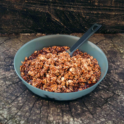 Porridge al cioccolato fondente Outfood in ciotola verde scuro con cucchiaio nero su tronco d'albero.