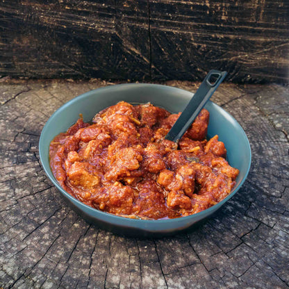 Spezzatino plant based Outfood alla Pizzaiola con pomodoro e origano in una ciotola blu scuro su un tronco di legno.