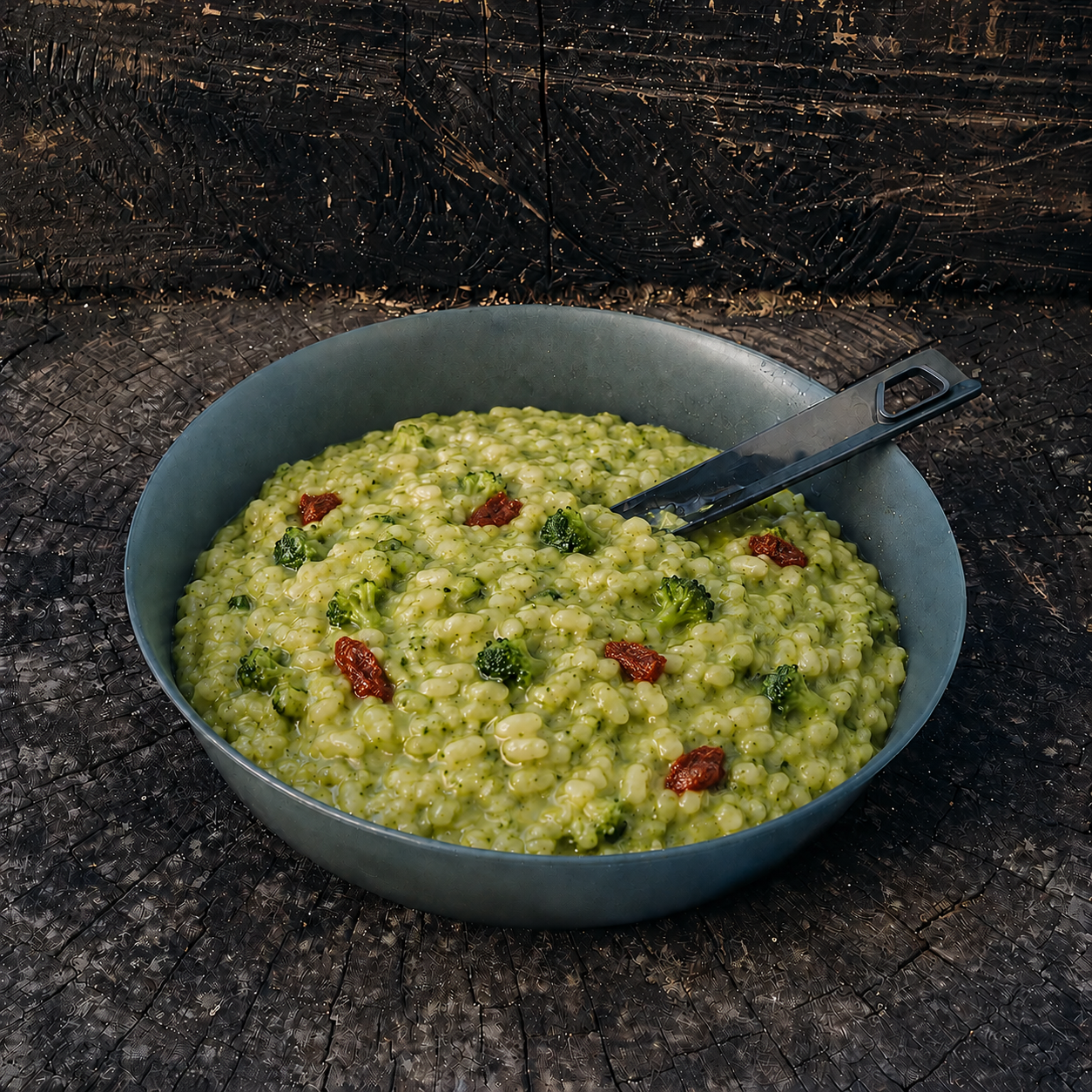 Risotto ai broccoli e pomodoro secco Outfood in ciotola grigia con cucchiaio nero su tronco di legno scuro.